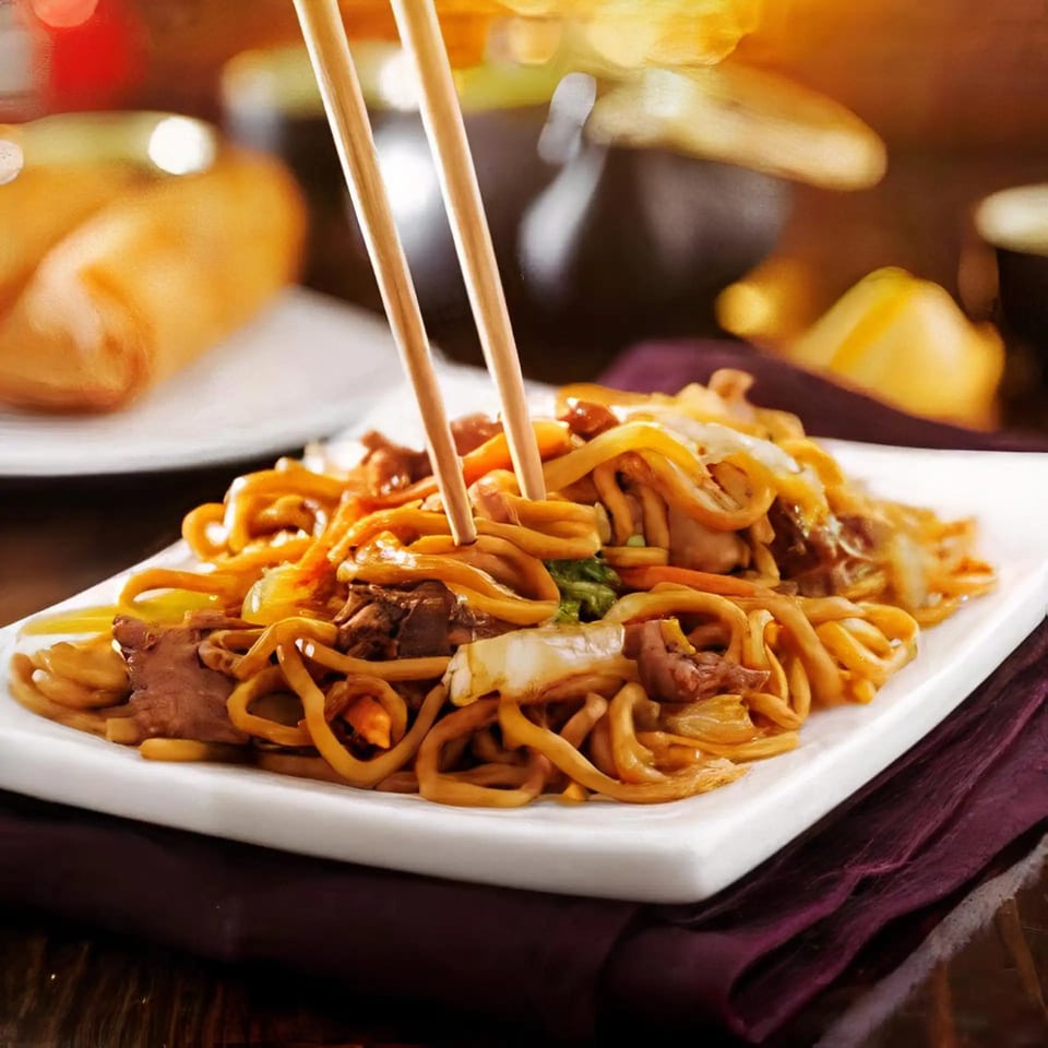 Chopsticks picking up a noodle from a plate of stir-fried noodles with meat and vegetables.