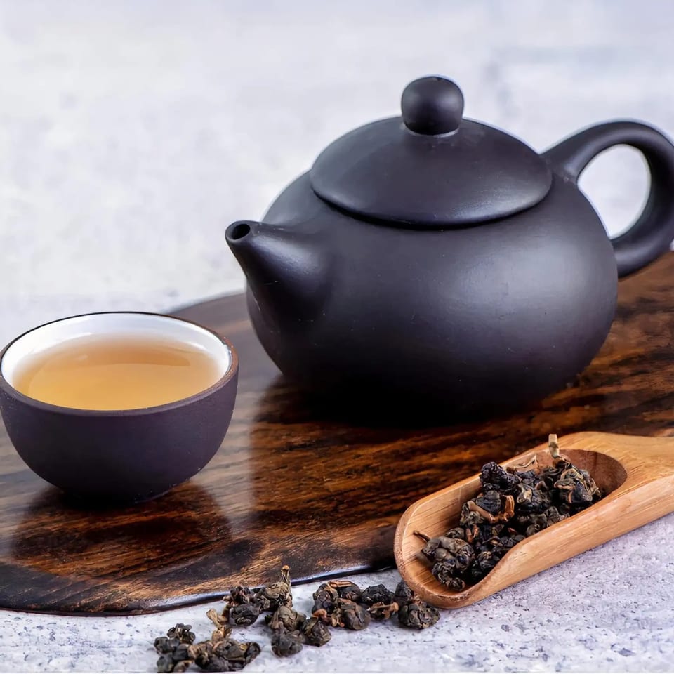 A black teapot and cup of tea sit on a wooden board, with loose tea leaves nearby.